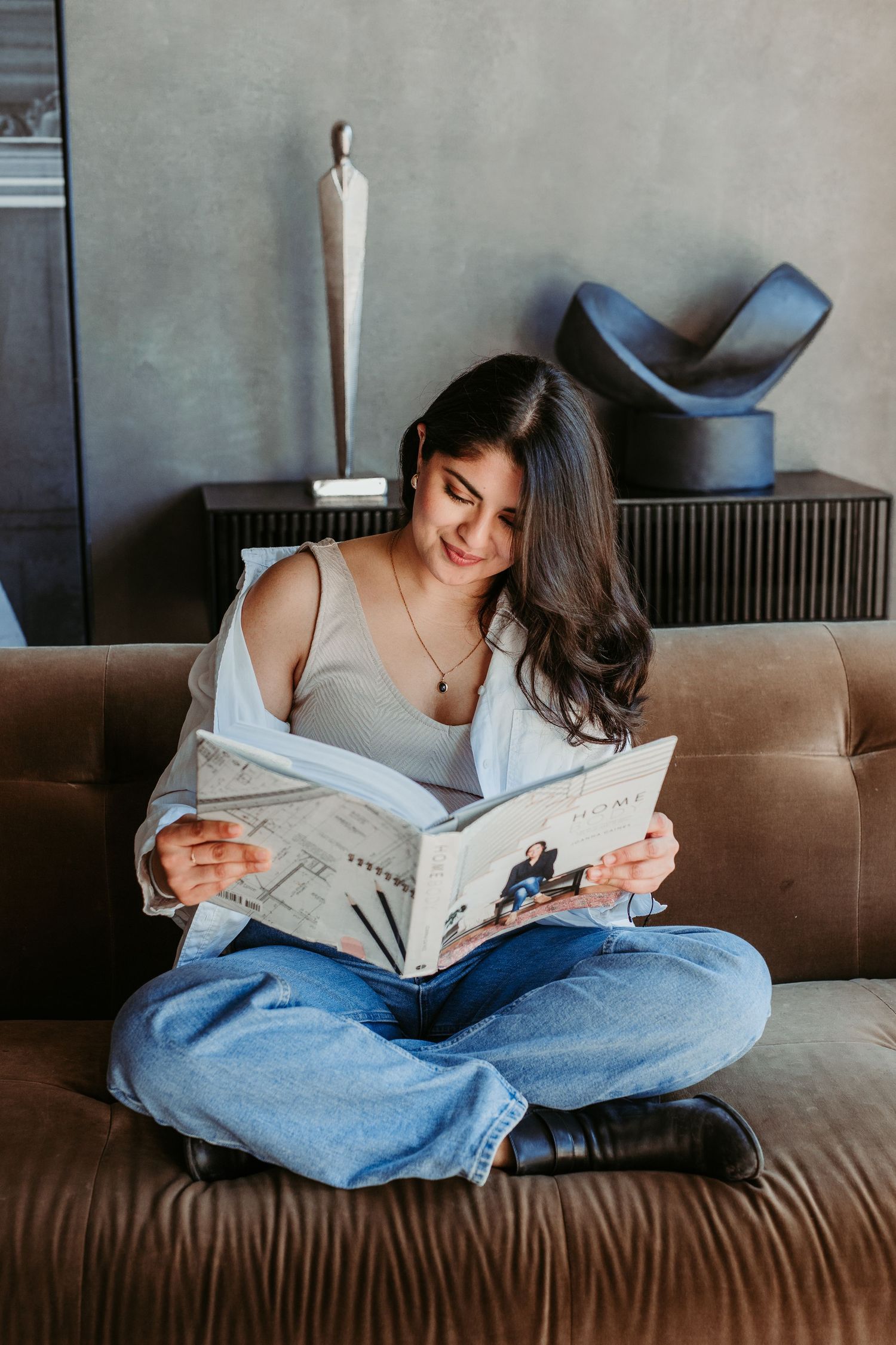 Person relaxing on brown couch while reading newspaper in casual white top and jeans in moody lighting.