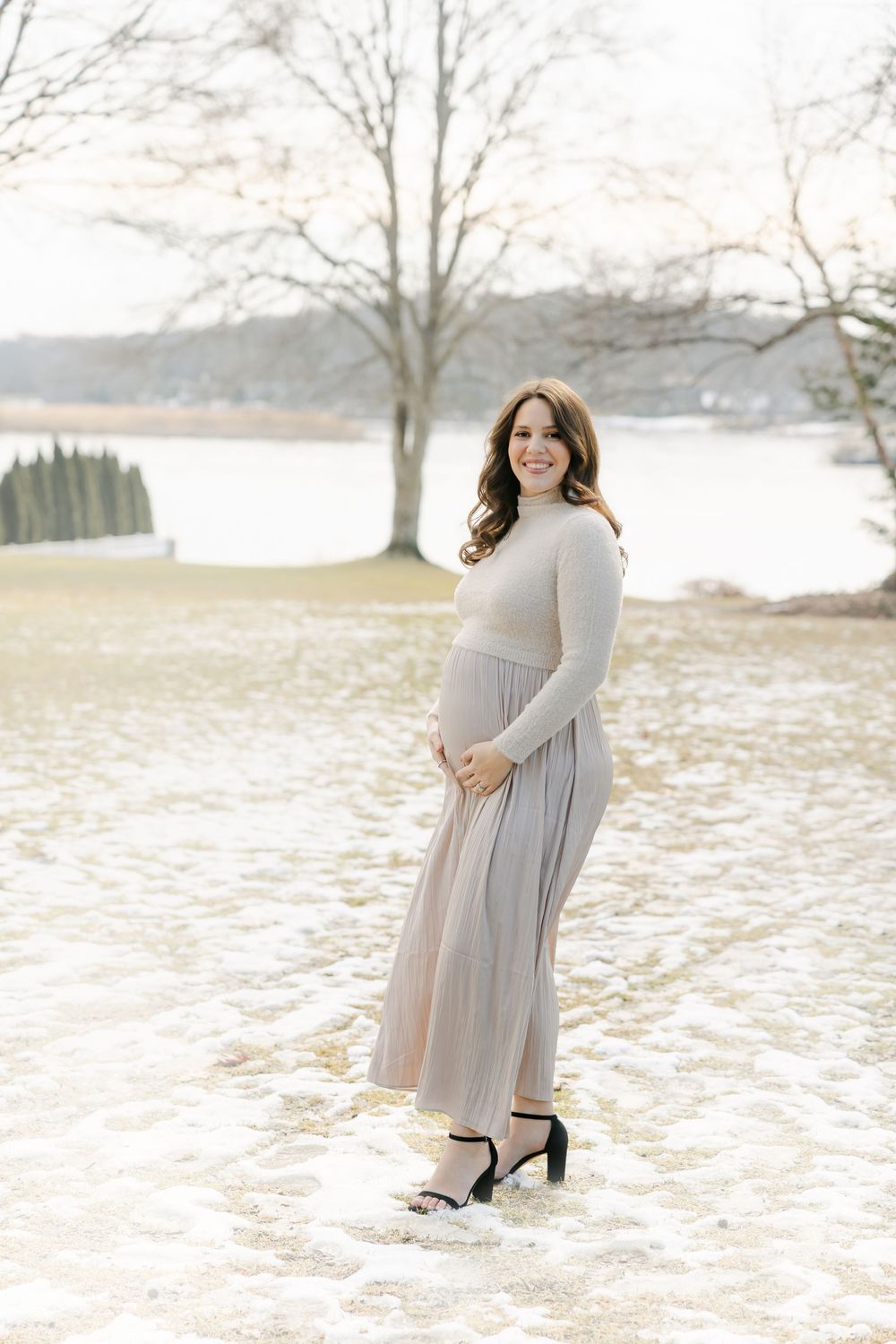 Winter maternity portraits on snowy ground with bare trees and water in the background. Hartford Photography