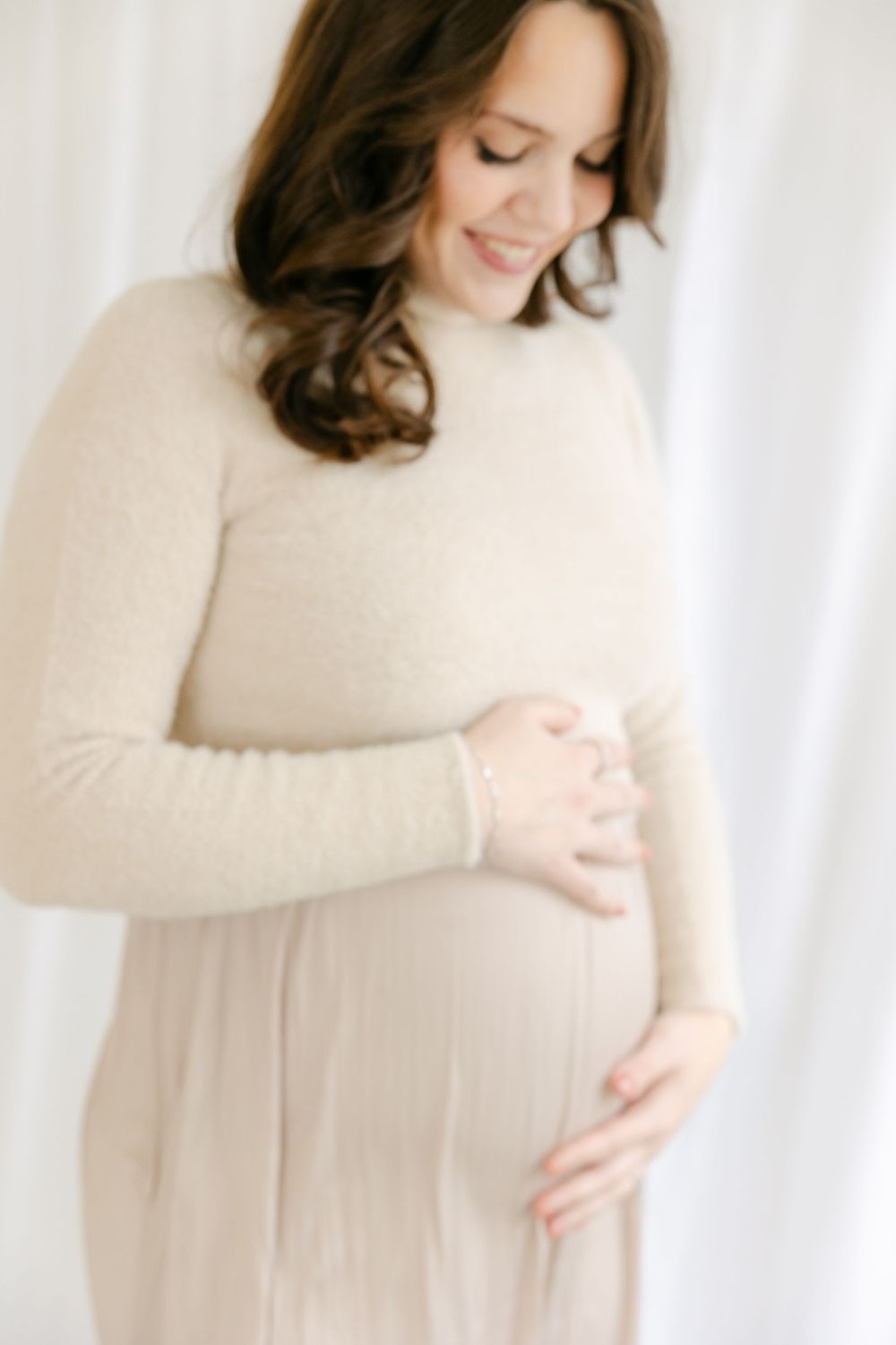 Expectant mother in cream sweater lovingly cradles her baby bump in soft natural light against white backdrop. Hartford Photography