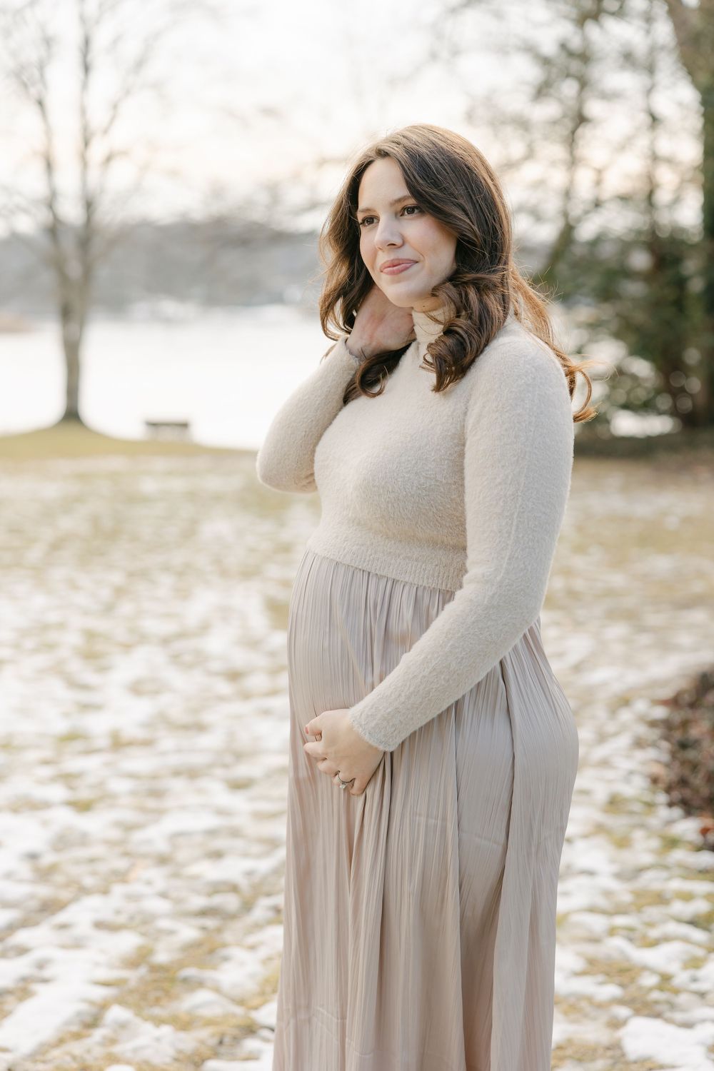 A maternity photo shoot in a snowy winter landscape with a woman wearing a cream-colored dress and sweater. Hartford Photography