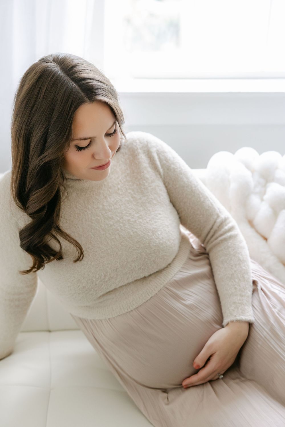 Maternity photoshoot featuring soft white bedding and a cozy cream sweater in natural light. Hartford Photography
