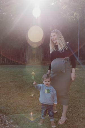 Outdoor maternity photo with sun flare and young child in casual wear.