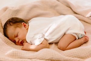Sleeping baby in white clothing curled up on beige bedding during naptime.