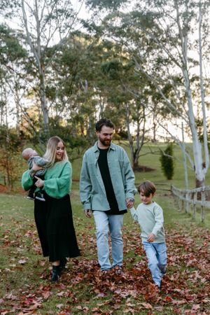 A family walks together through fallen autumn leaves in a park on a sunny afternoon.