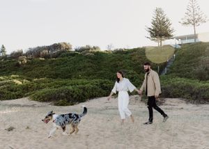 A couple walks their spotted dog along a coastal beach path at sunset.
