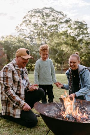 Family gathering around a fire pit in an outdoor setting with autumn trees in background.