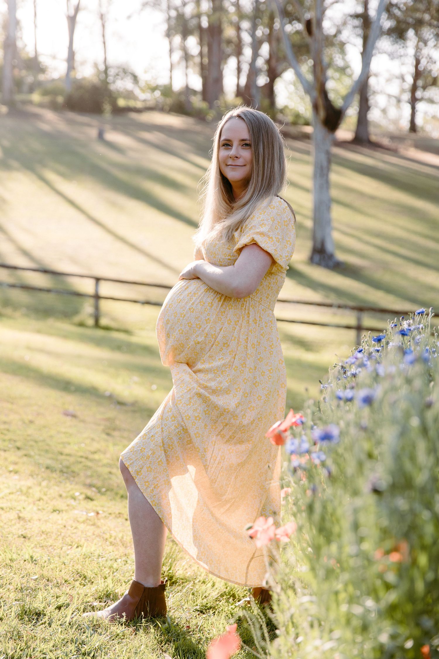 Expectant mother in yellow dress poses in sunlit garden with spring flowers.