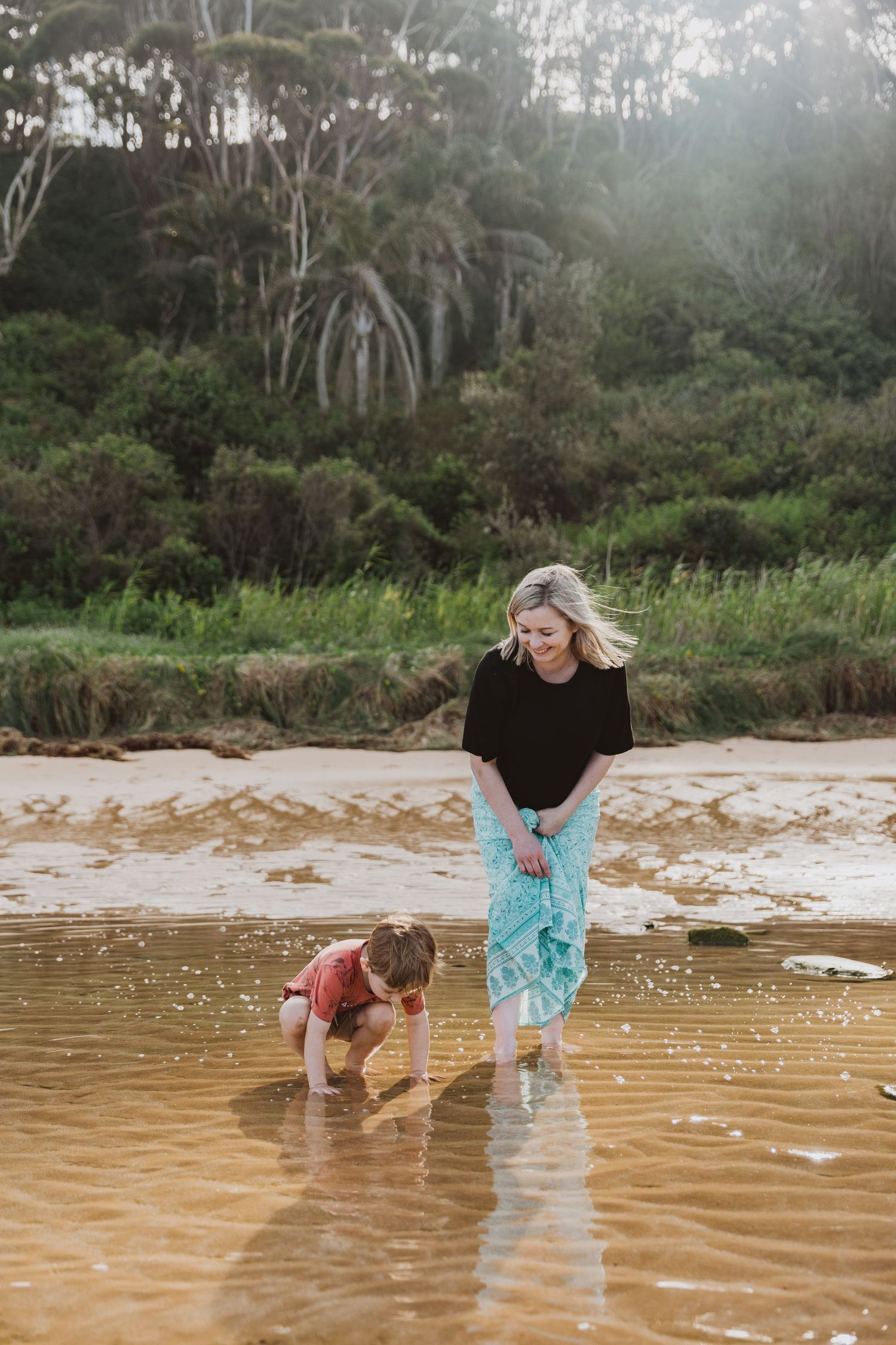 Two people playing and exploring in shallow water at a coastal beach with trees in background.