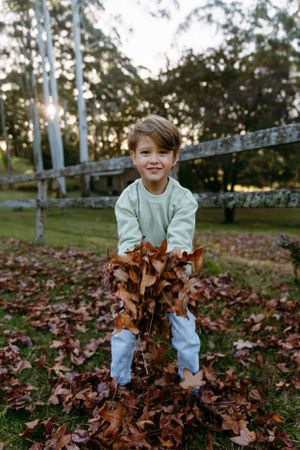 Child in light sweater plays joyfully with pile of autumn leaves in backyard.