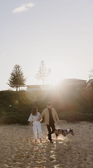 A couple holding hands walks their dog on a sandy beach at golden hour.