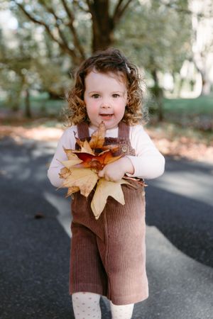 Young child in brown overalls holding autumn leaves on a path.