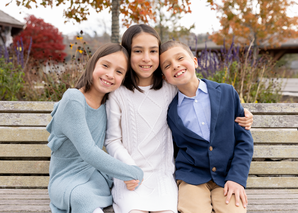 Three siblings smile and embrace while seated on a bench with fall trees behind them.
