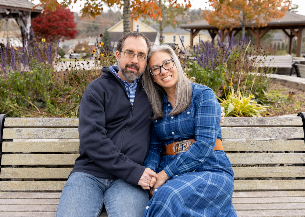 A couple sits closely together on a bench, smiling warmly in an autumn garden setting.