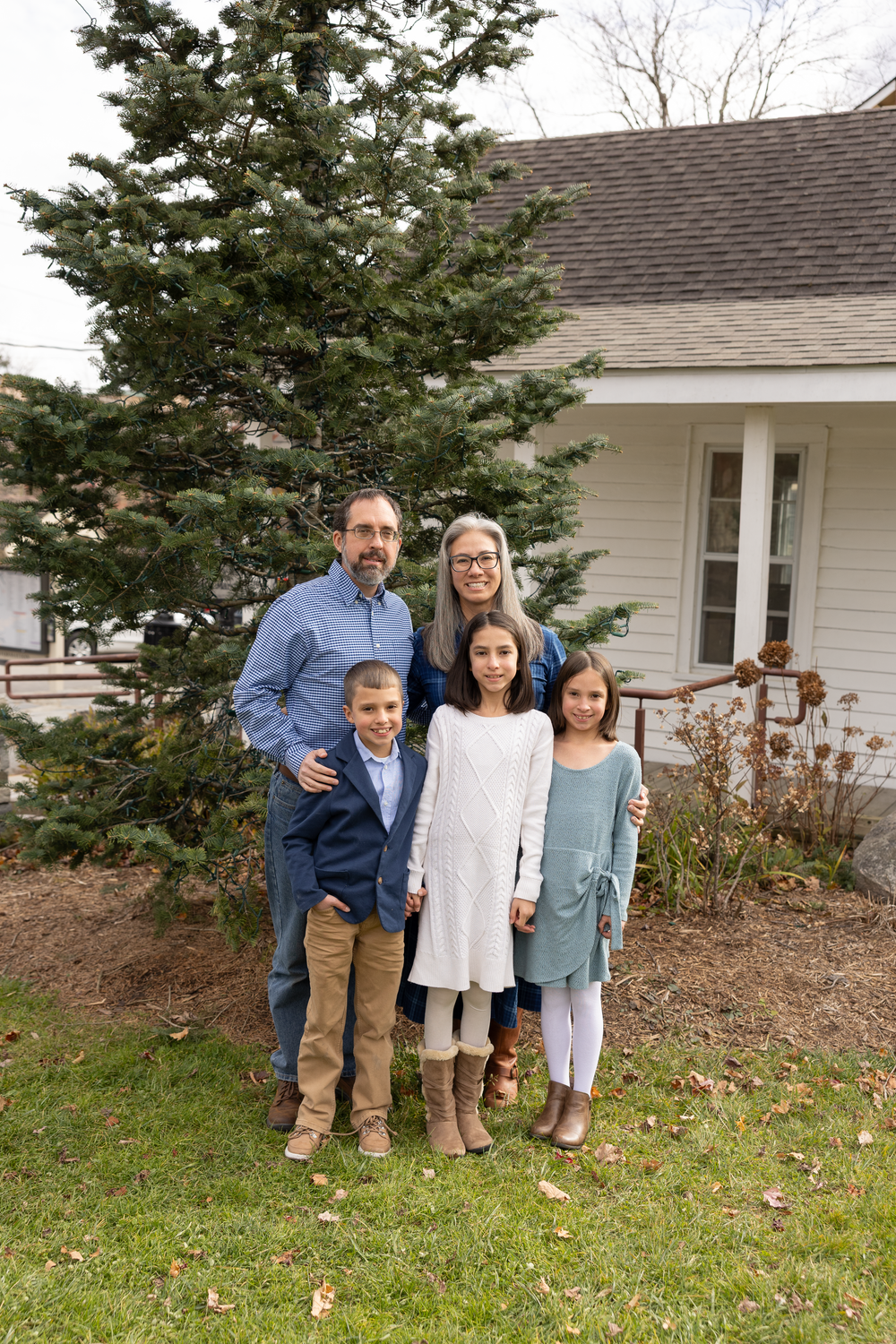 A family of five poses together on a lawn beside a large evergreen tree and white home.