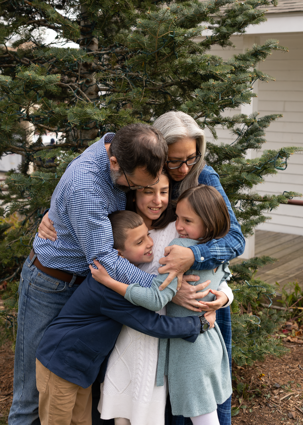 A family of five shares a joyful group hug near a tall evergreen tree outdoors.