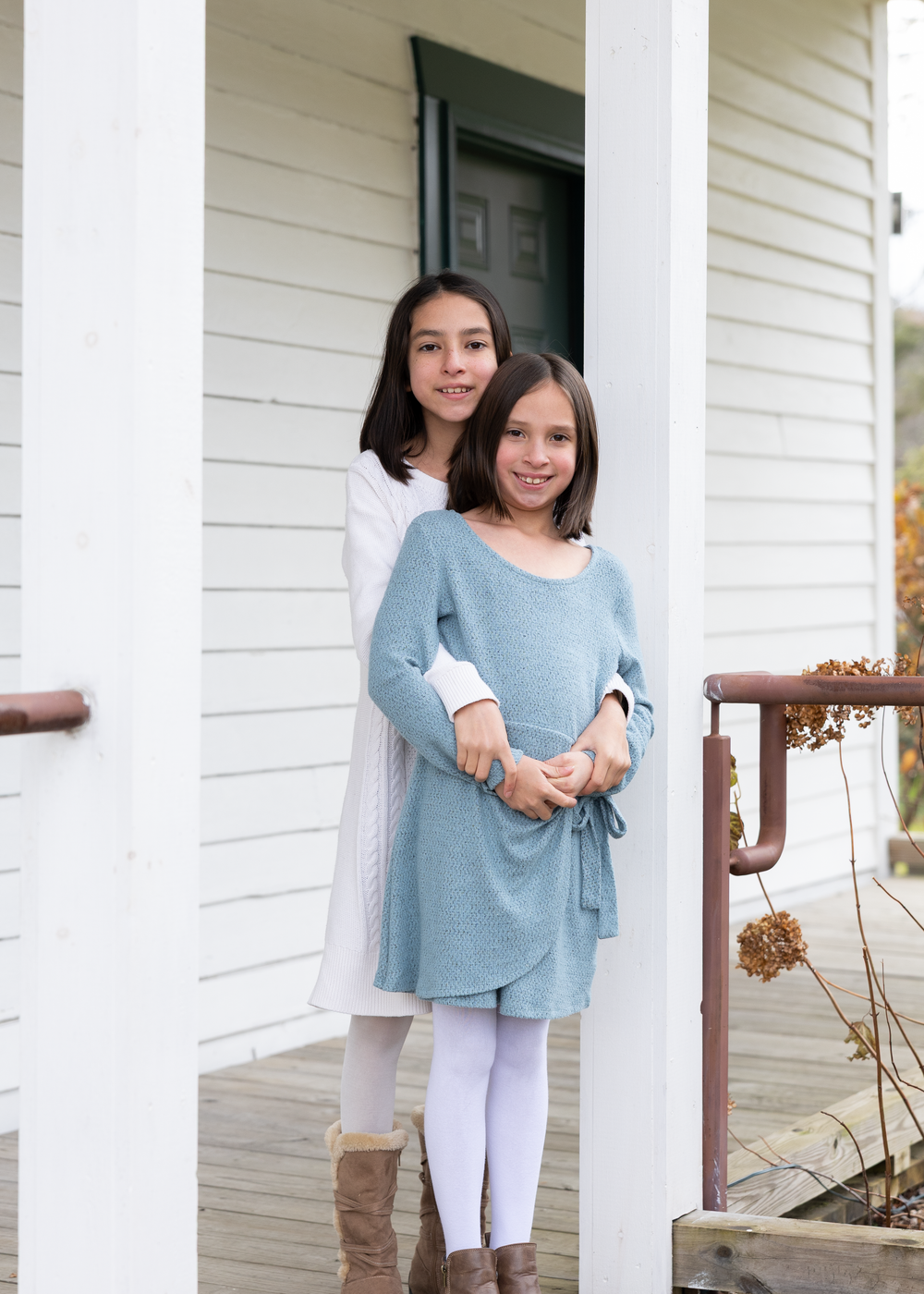 Two sisters stand together on a white porch, smiling warmly in coordinating outfits.