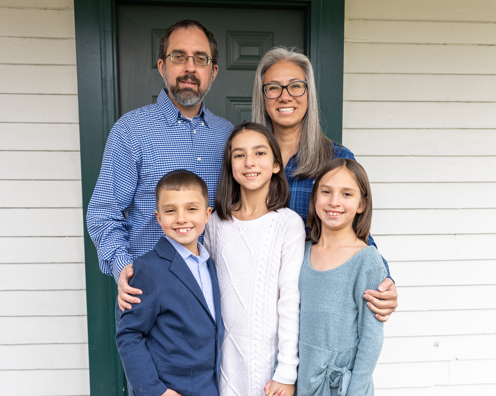 Family of five smiling together on a porch in front of a green door, dressed in coordinated outfits.