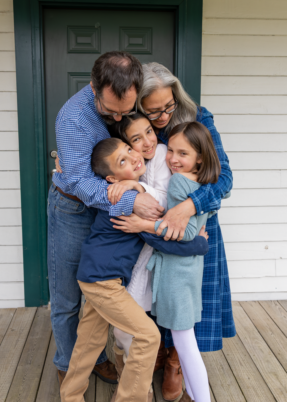 Loving family of five sharing a warm group hug on a wooden porch outside their home.