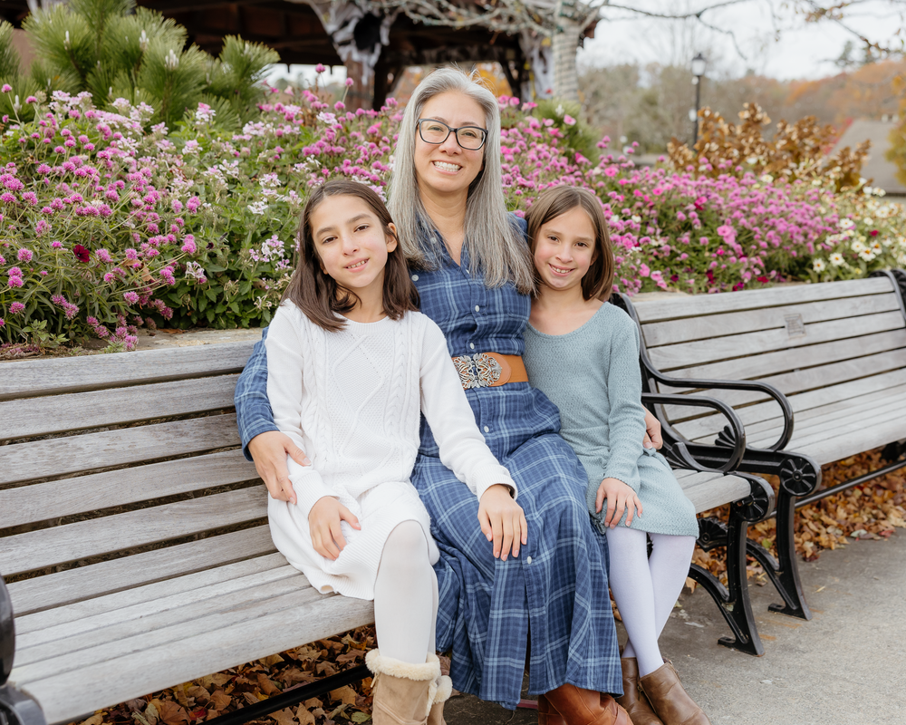 Woman with gray hair and glasses sitting on a bench with two young girls amid pink flowers.