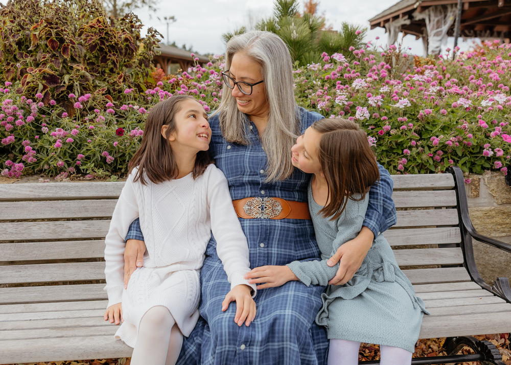 Mother with gray hair smiling at two daughters on a garden bench surrounded by pink blooms.