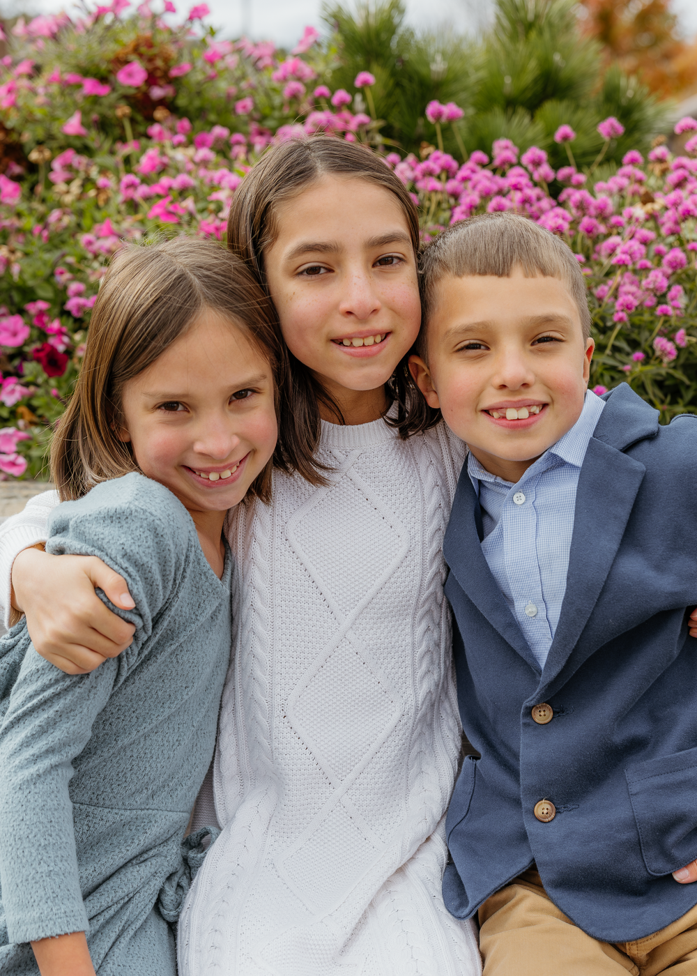 Three siblings hugging and smiling together in front of vibrant pink flowers outdoors.