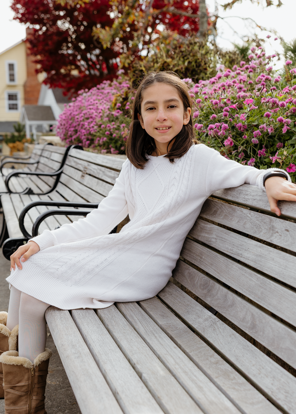 Young girl in white dress relaxing on a wooden bench with colorful flowers in the background.