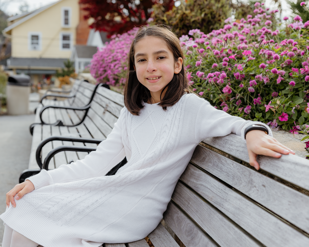 Smiling young girl in a white dress sitting on a bench near bright pink garden flowers.