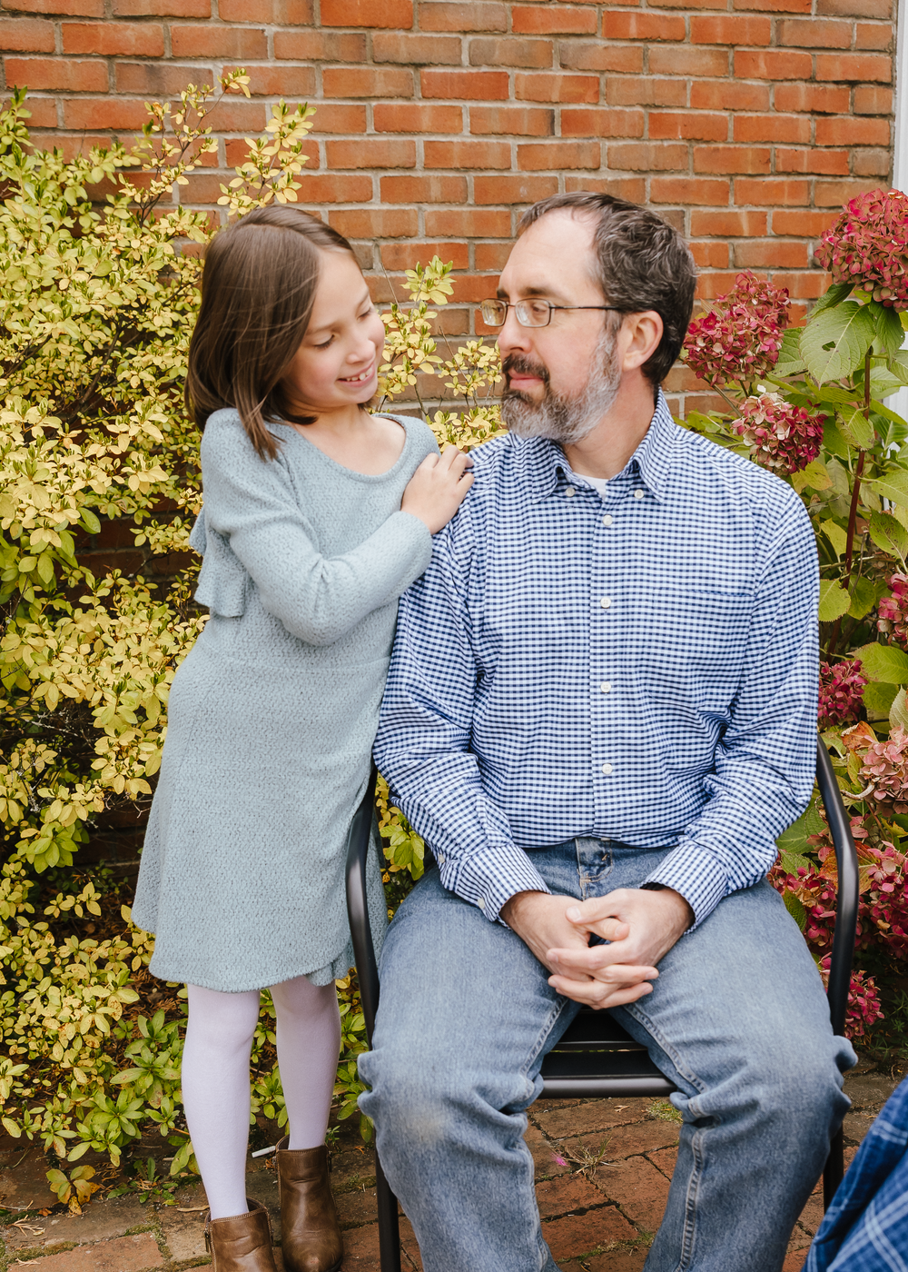 Young girl in gray dress talking to her father seated outdoors near a brick wall and greenery.