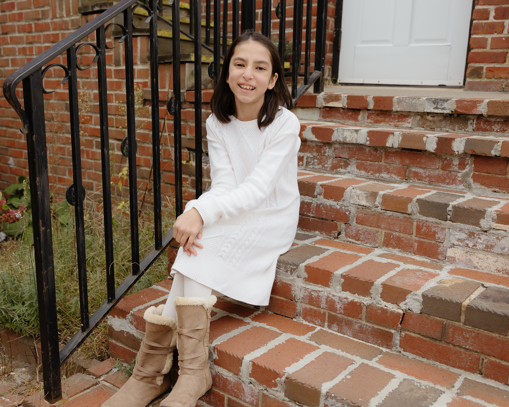 Young girl in white dress smiling while sitting on brick steps beside a black iron railing.