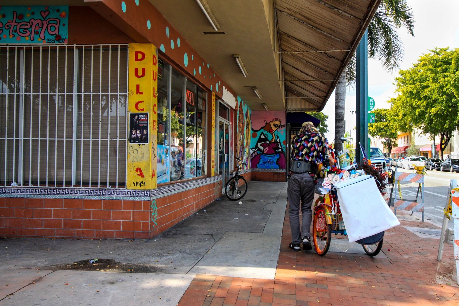 People walking past small retail shops with security bars on a sunny day in a downtown shopping district.