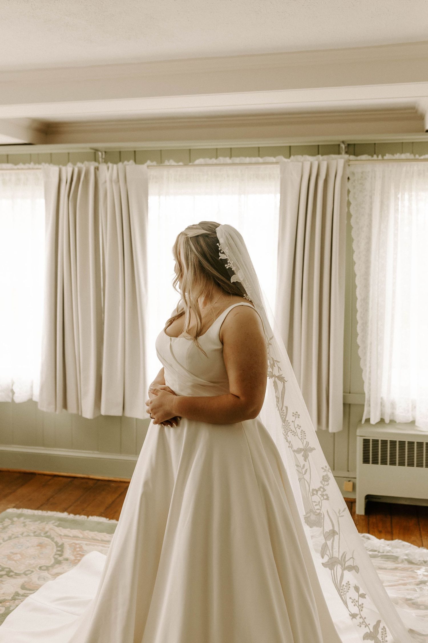 Bride in satin gown with embroidered cathedral veil standing by window in green room.