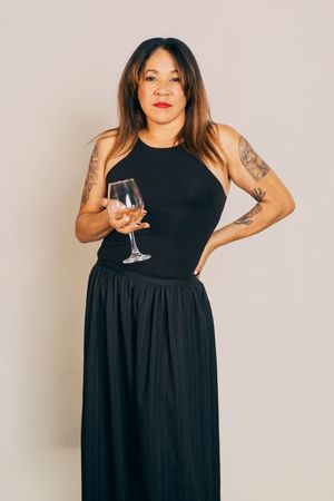 Woman in black maxi dress posing with wine glass against white background.