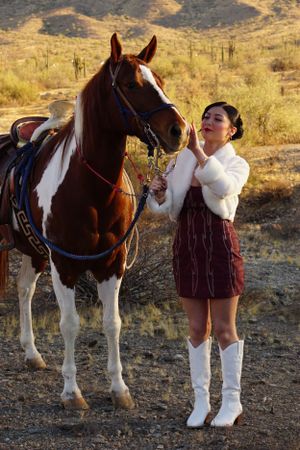 Person posing playfully with a brown and white horse in an arid desert landscape.