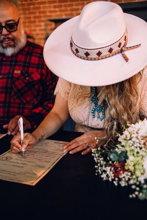 A person in a white hat signs a document with flowers nearby on a wooden surface.