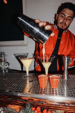 Bartender pouring multiple cocktails on metallic bar counter.