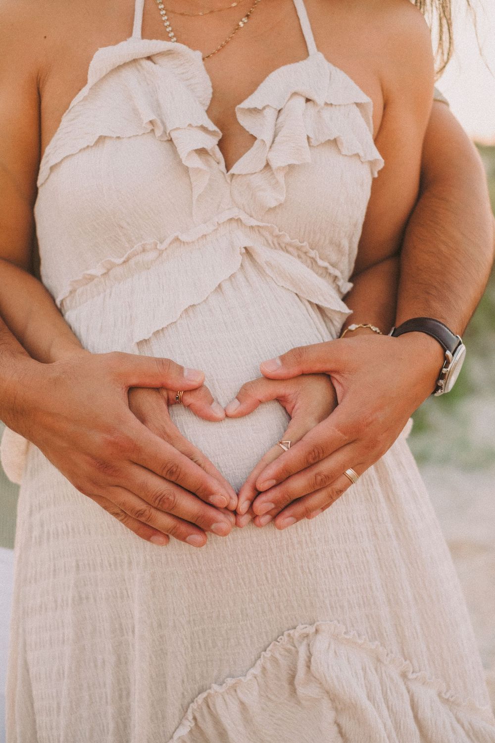Couple forms heart shape with hands on pregnant belly in white ruffled maternity dress.