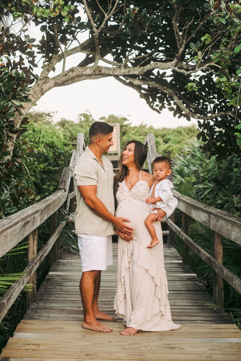 Family of three poses on wooden bridge surrounded by lush tropical greenery at beach.
