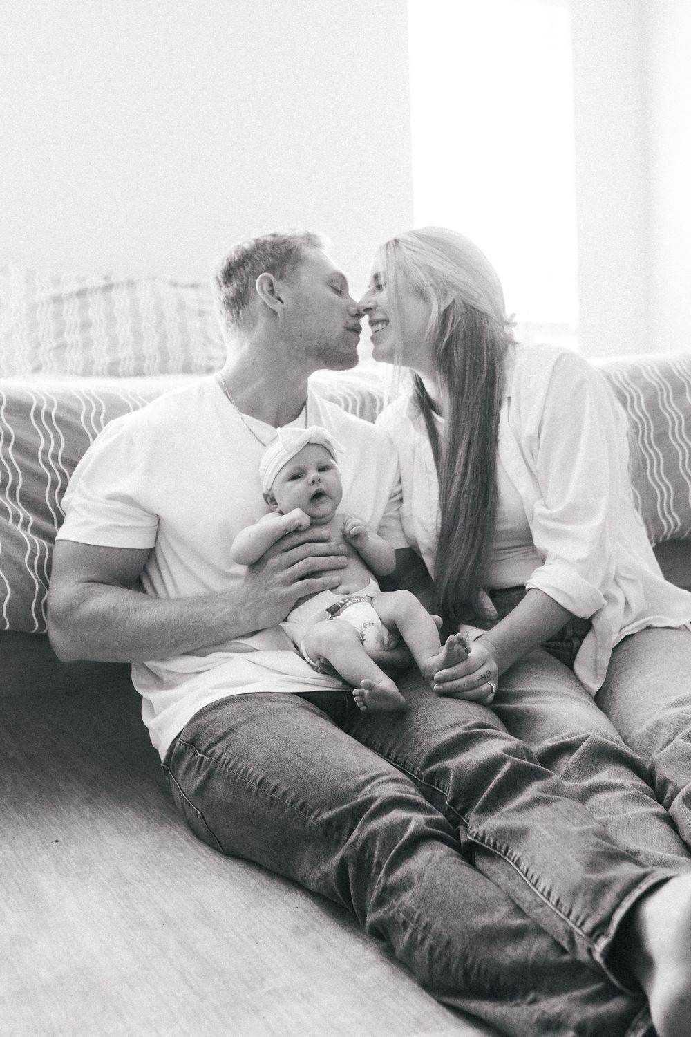 Black and white photo of parents sharing a kiss while holding their smiling baby on living room floor.