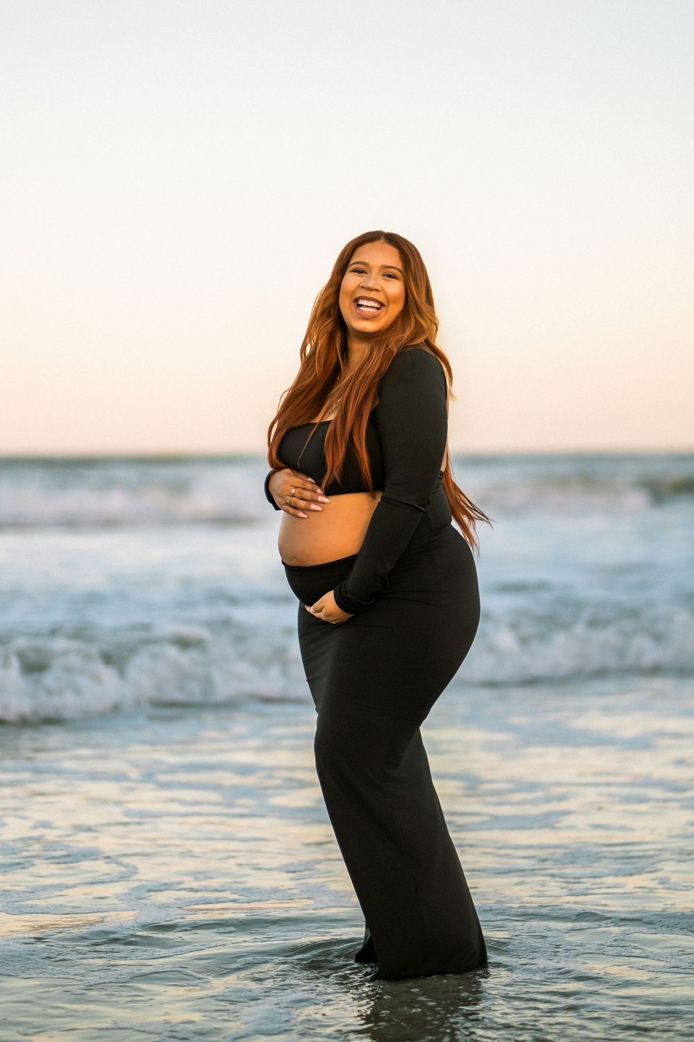 Laughing pregnant woman in black crop top and pants standing in ocean waves at sunset, holding baby bump.