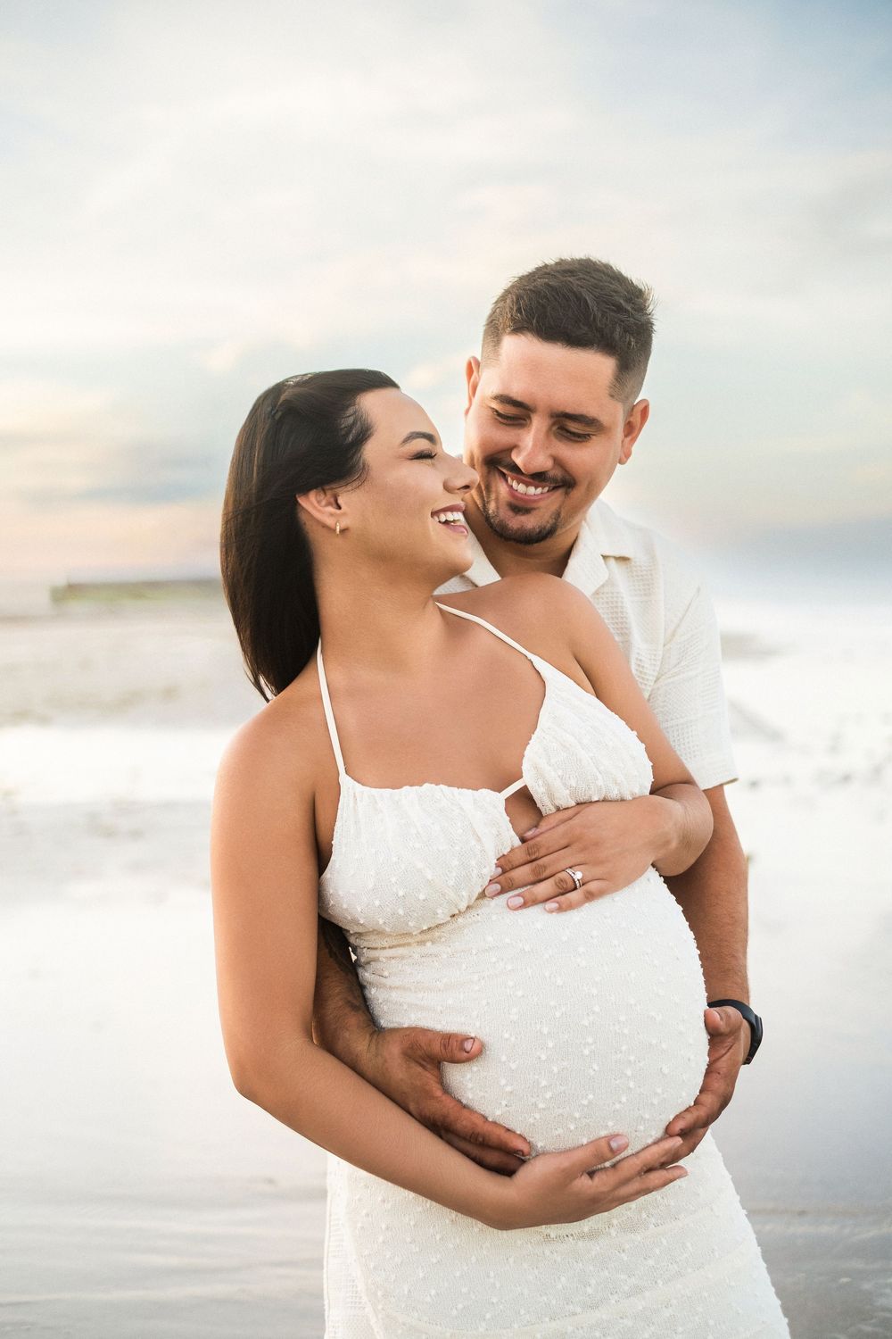 A smiling couple in white outfits embraces on a beach, with the man tenderly holding the pregnant woman's belly.
