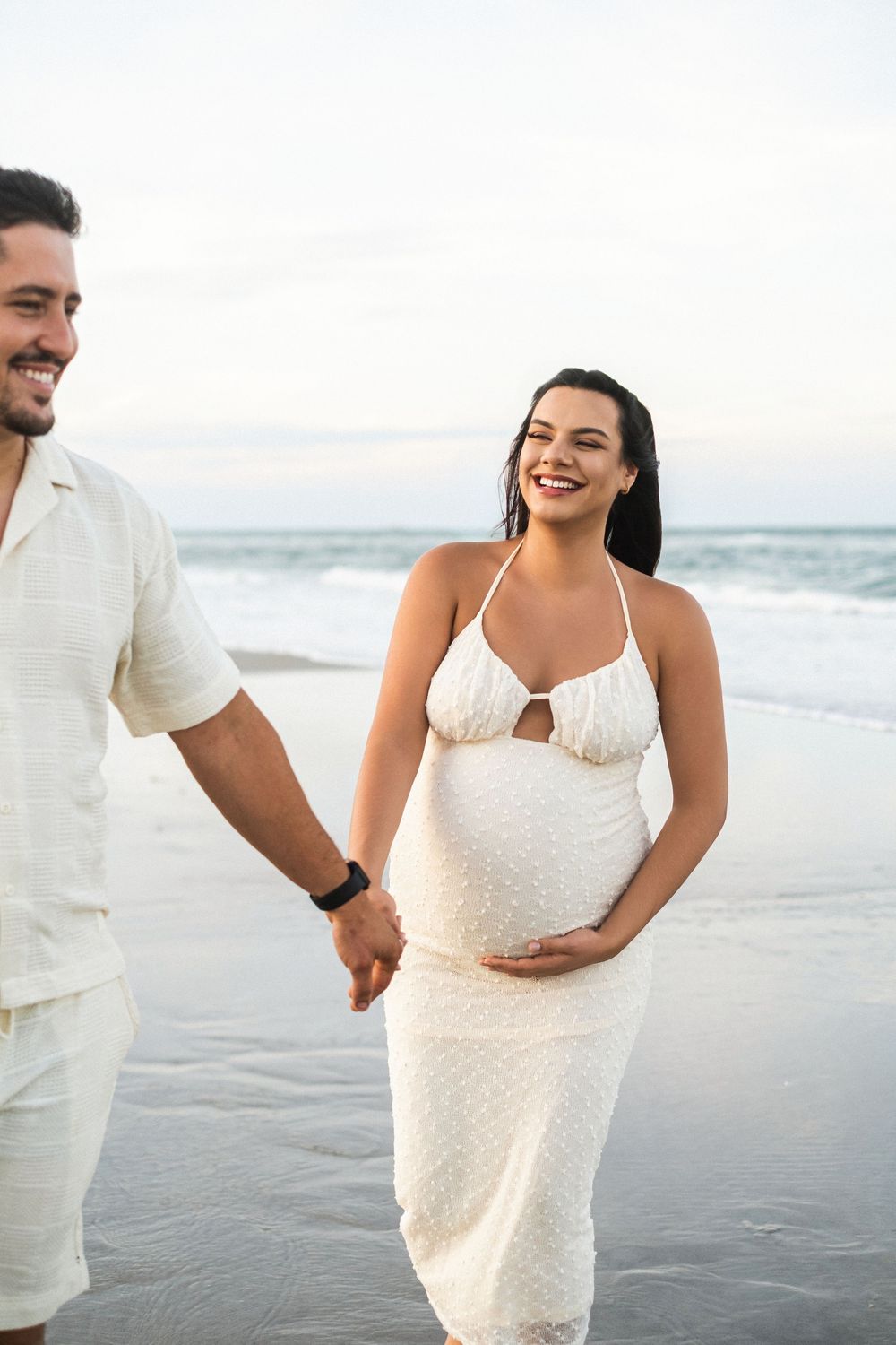 Smiling pregnant woman in white halter dress holds hands with man on beach at sunset.