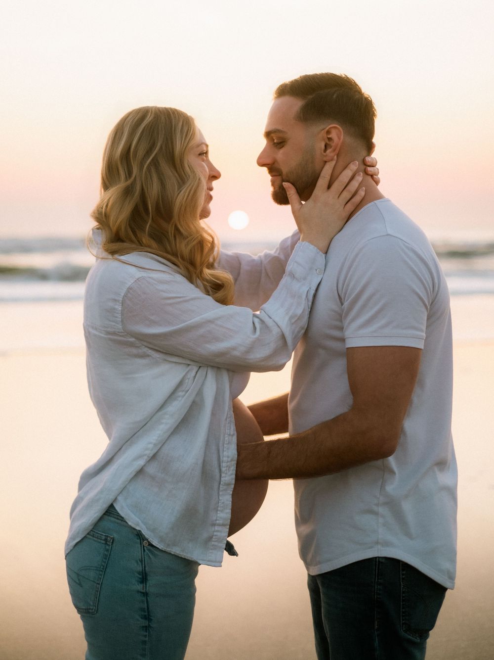 Loving couple facing each other at sunset beach, woman gently holding mans face near ocean waves.