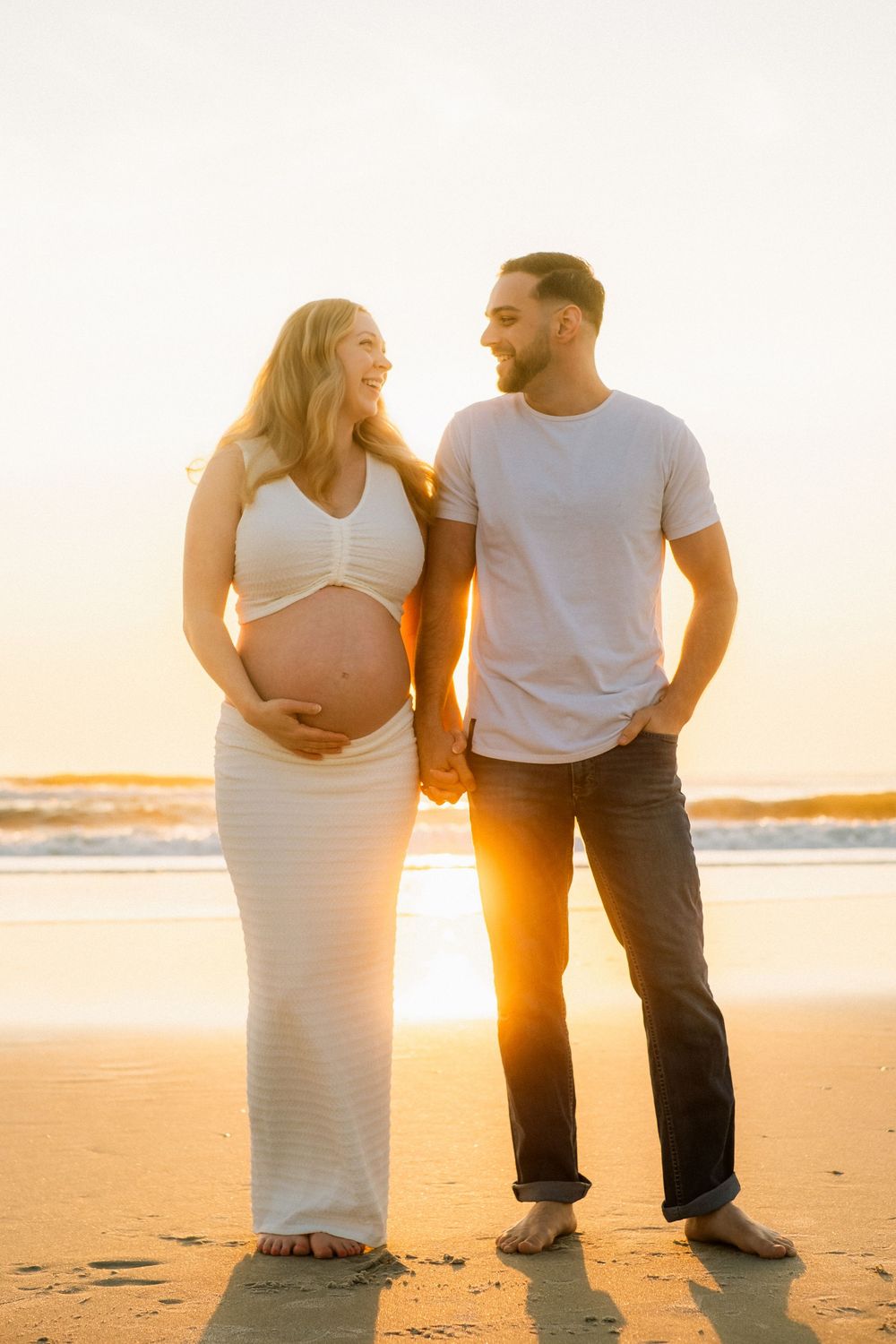 Smiling pregnant couple holding hands on beach at sunset, woman in white dress touching baby bump.