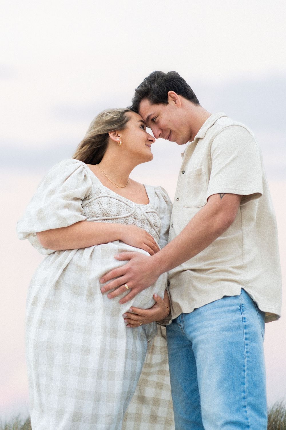Smiling couple touching foreheads tenderly while both cradling pregnant belly outdoors at dusk.
