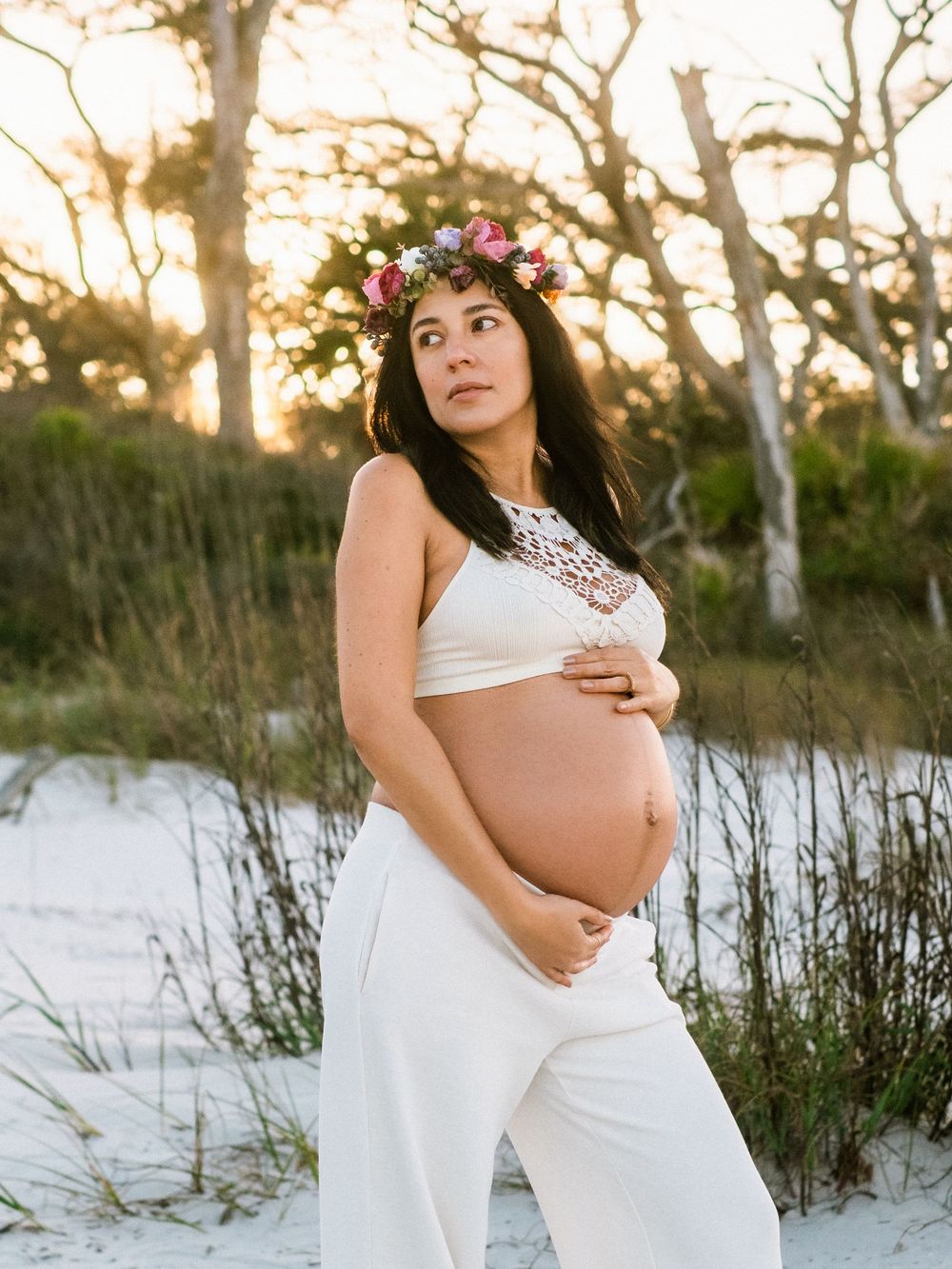 Pregnant woman in white crop top and pants with floral crown poses on sandy beach at sunset.