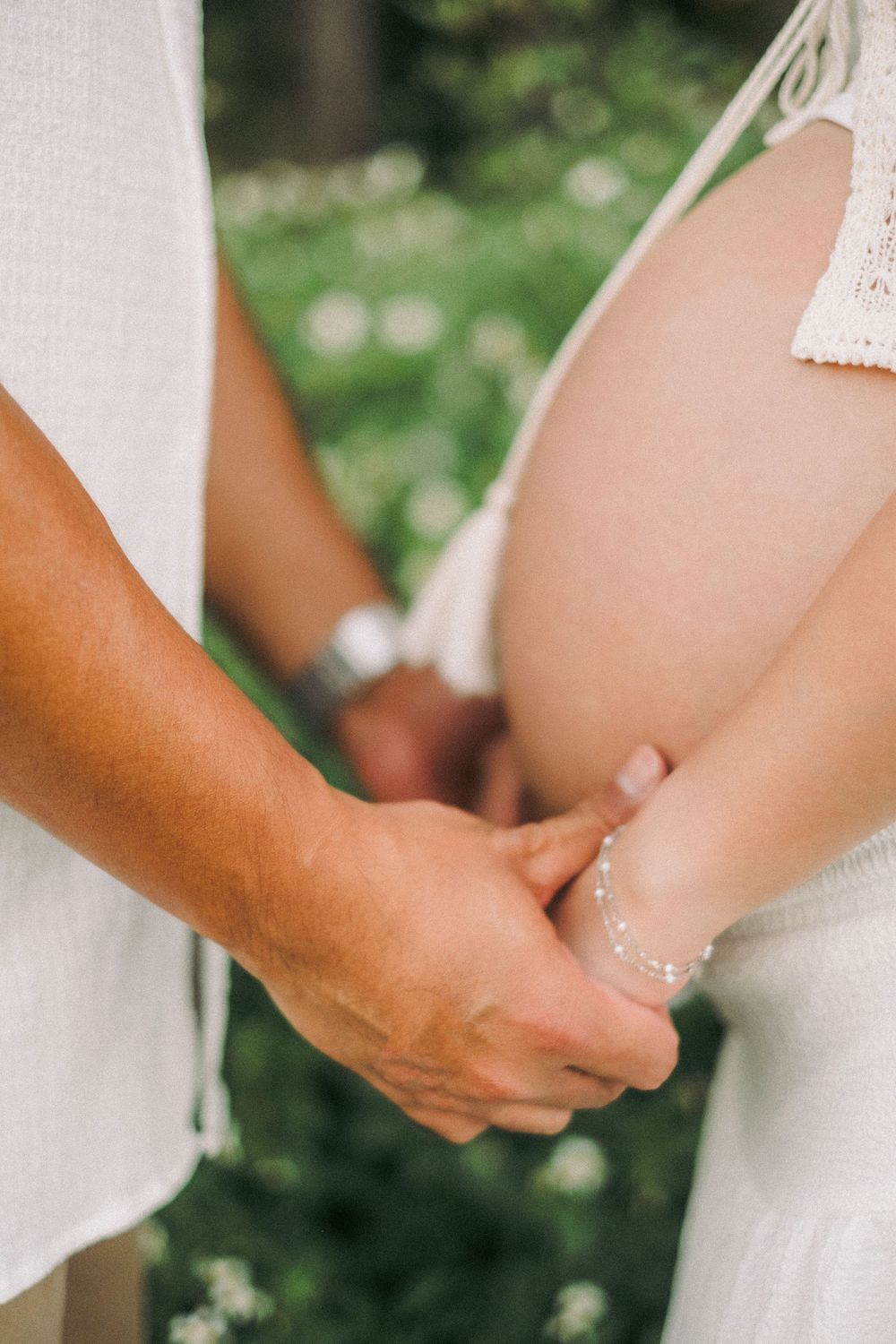 Close-up of couple holding hands showing pregnant belly with pearl bracelet outdoors.