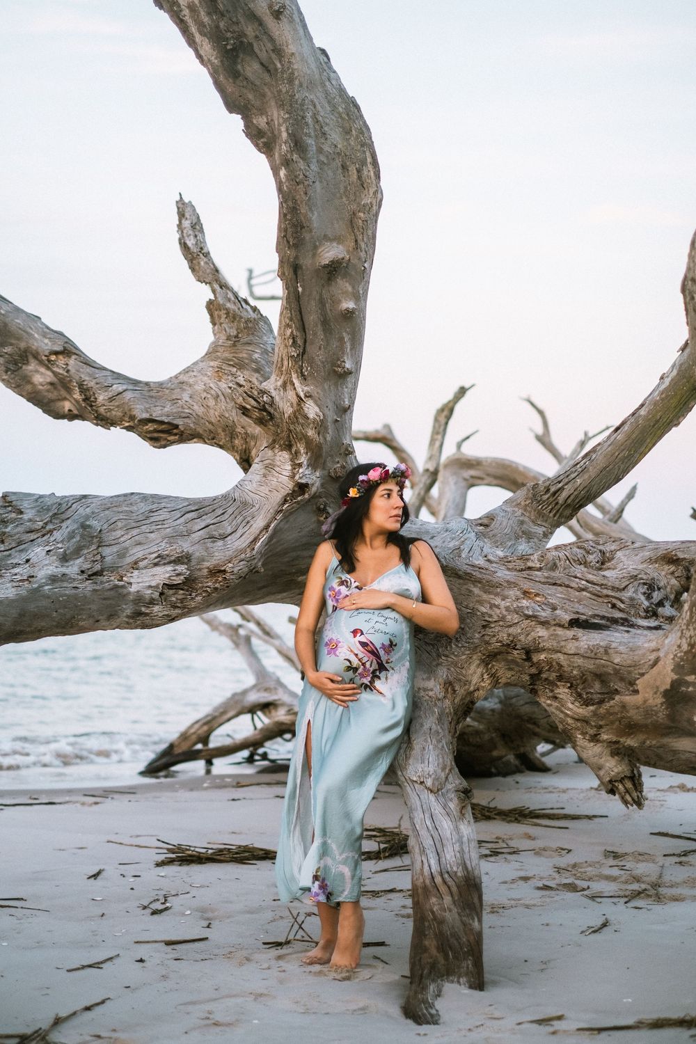 Pregnant woman in mint floral dress with flower crown leans against large driftwood by ocean.