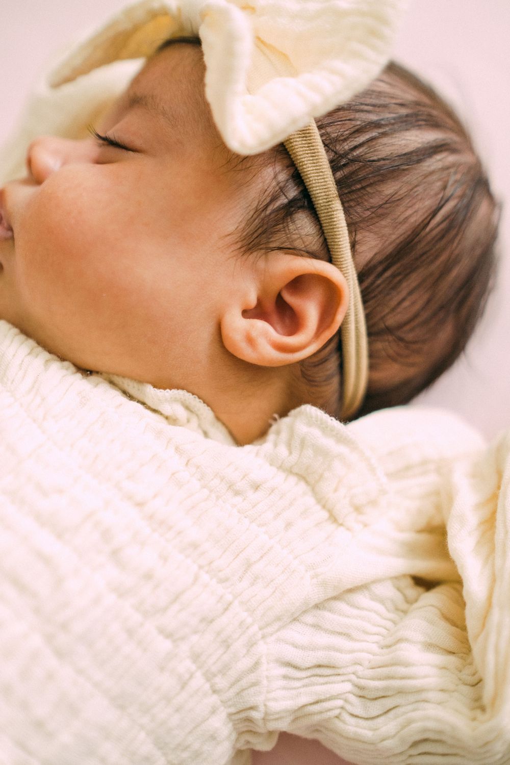 Close-up of peaceful sleeping newborn baby with dark hair wearing cream bow headband.