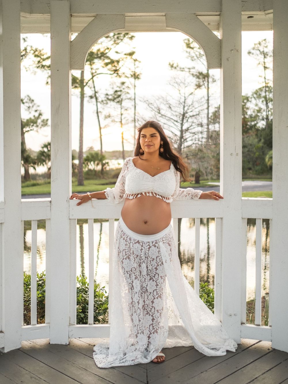 Pregnant woman in white lace maternity gown posing in a white gazebo at sunset.