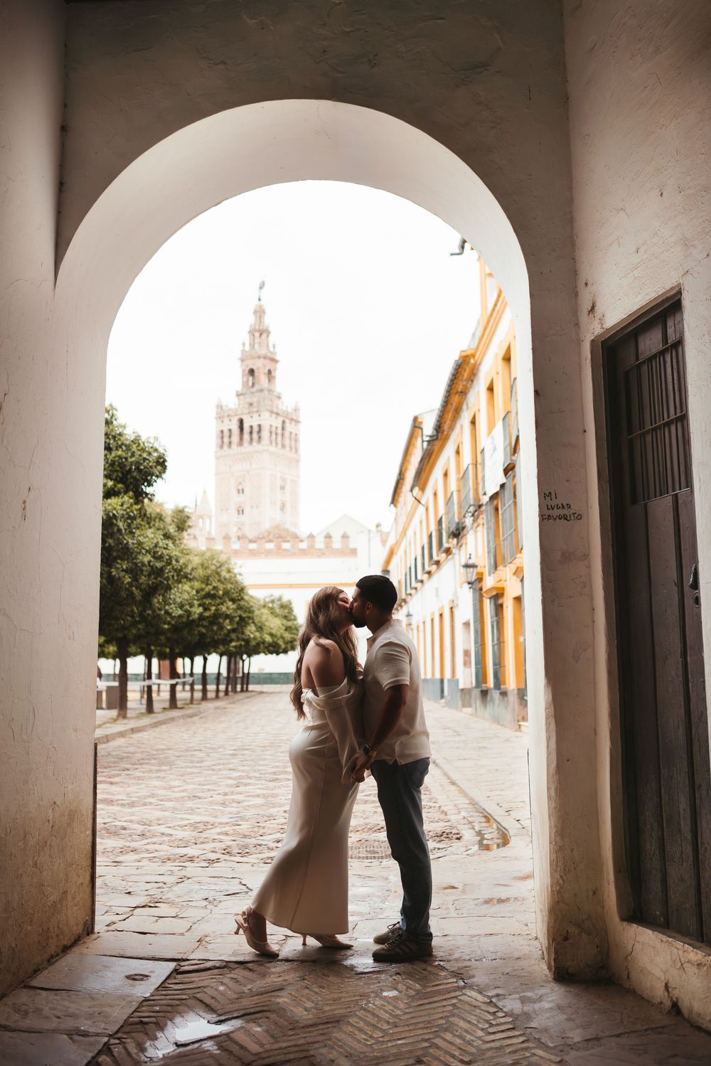 A romantic wedding couple shares intimate moments under white arched colonnade walkways with a church tower visible in distance.